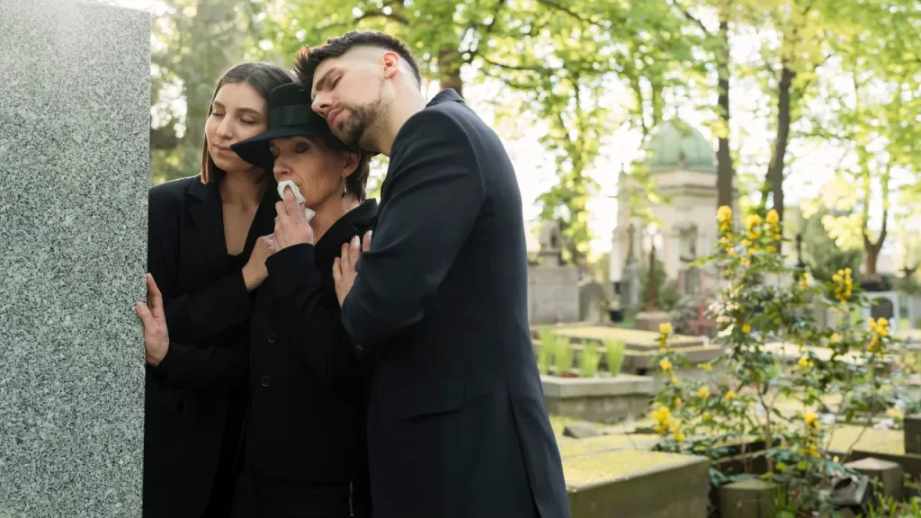 Família vestida de preto se abraça emocionada diante de um túmulo em um cemitério arborizado durante um funeral.