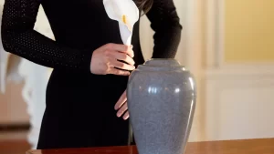 Pessoa segurando uma flor branca ao lado de urna cinza durante cerimônia de cremação.